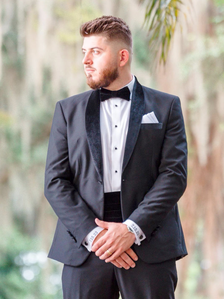 Man wearing a black velvet shawl collar tuxedo made of 100% Italian wool, standing outdoors.