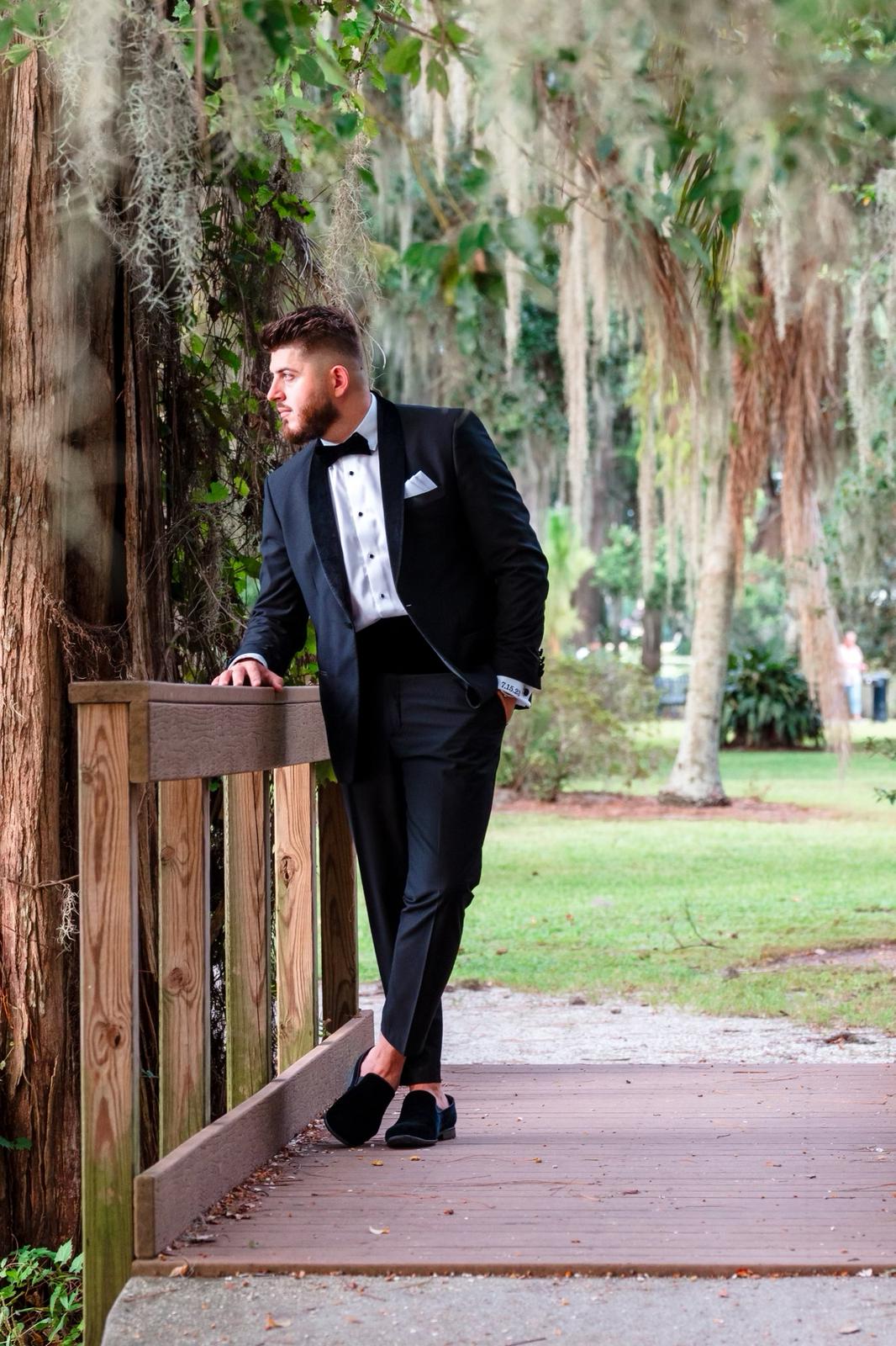 Man wearing a velvet shawl lapel tuxedo made from 100% Italian wool, standing on a wooden bridge in a lush park setting.