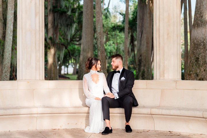 Bride in long-sleeve lace gown and groom in tuxedo sit on stone bench in a wooded area, sharing a loving look.